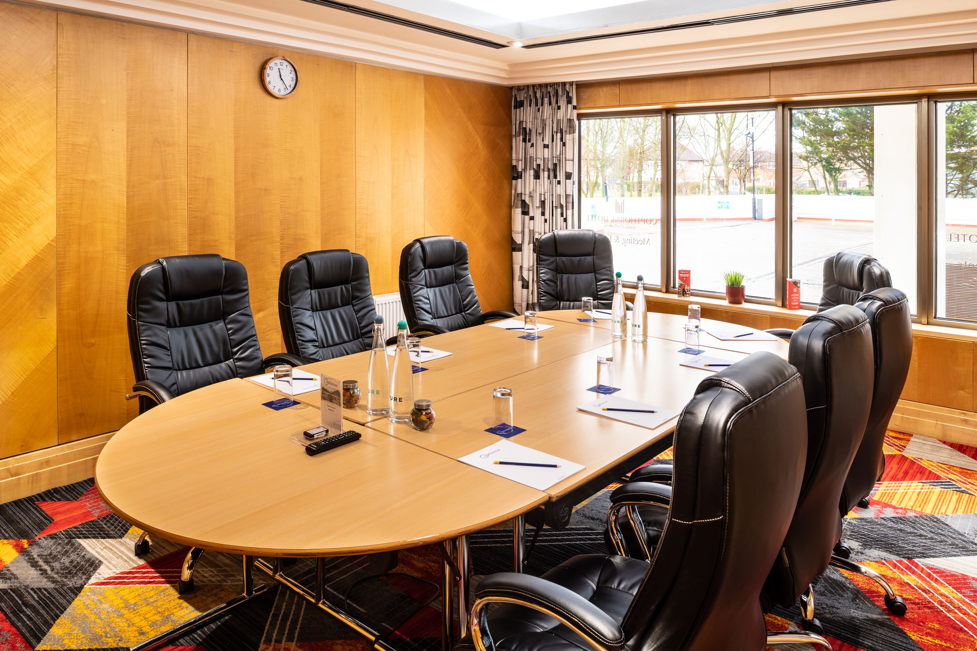 Un salón de conferencias con una mesa de madera, sillas de cuero negro, un reloj en la pared y una ventana con vistas al exterior.