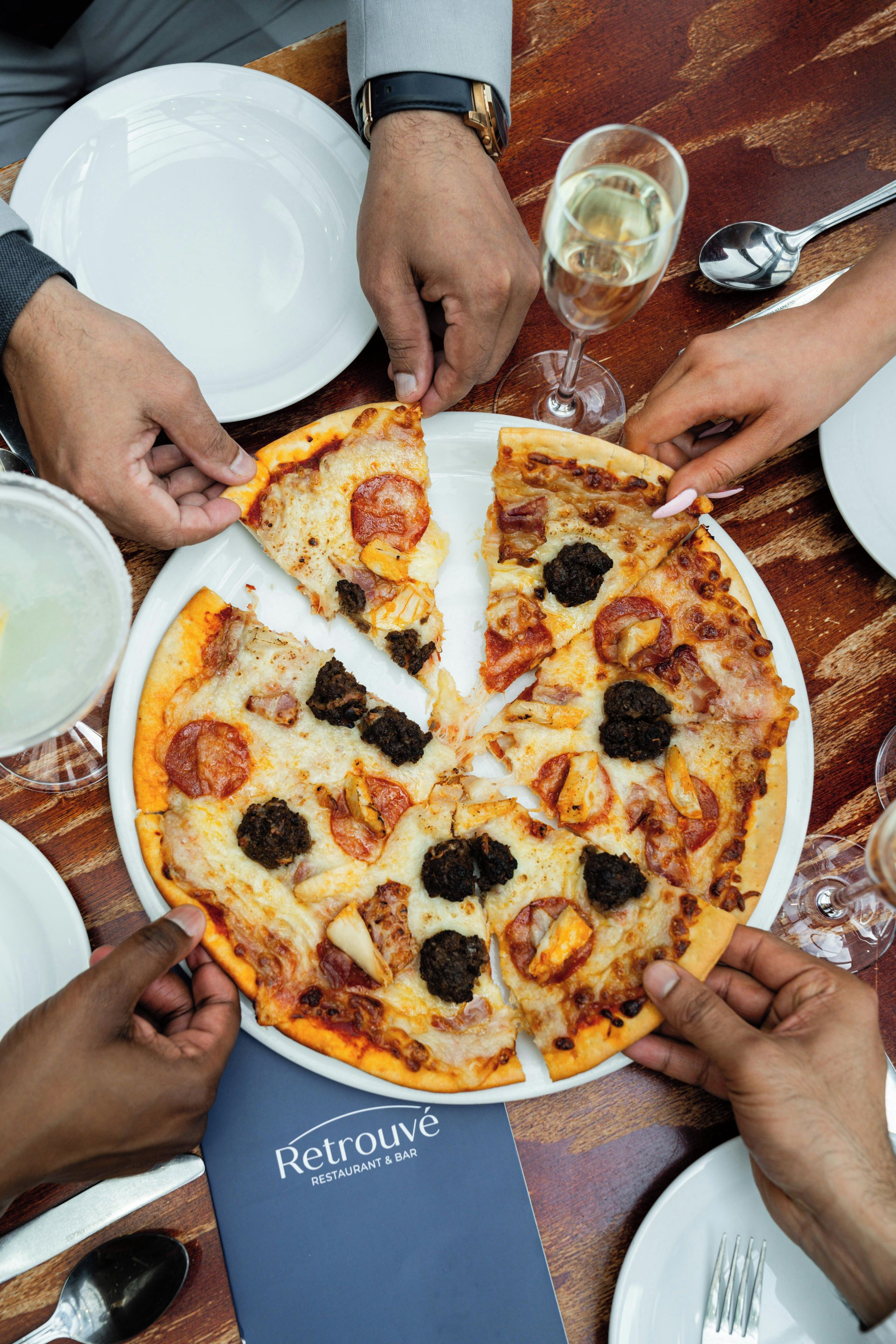 Una pizza con diversi condimenti, un bicchiere di vino bianco e un cucchiaio su un tavolo.