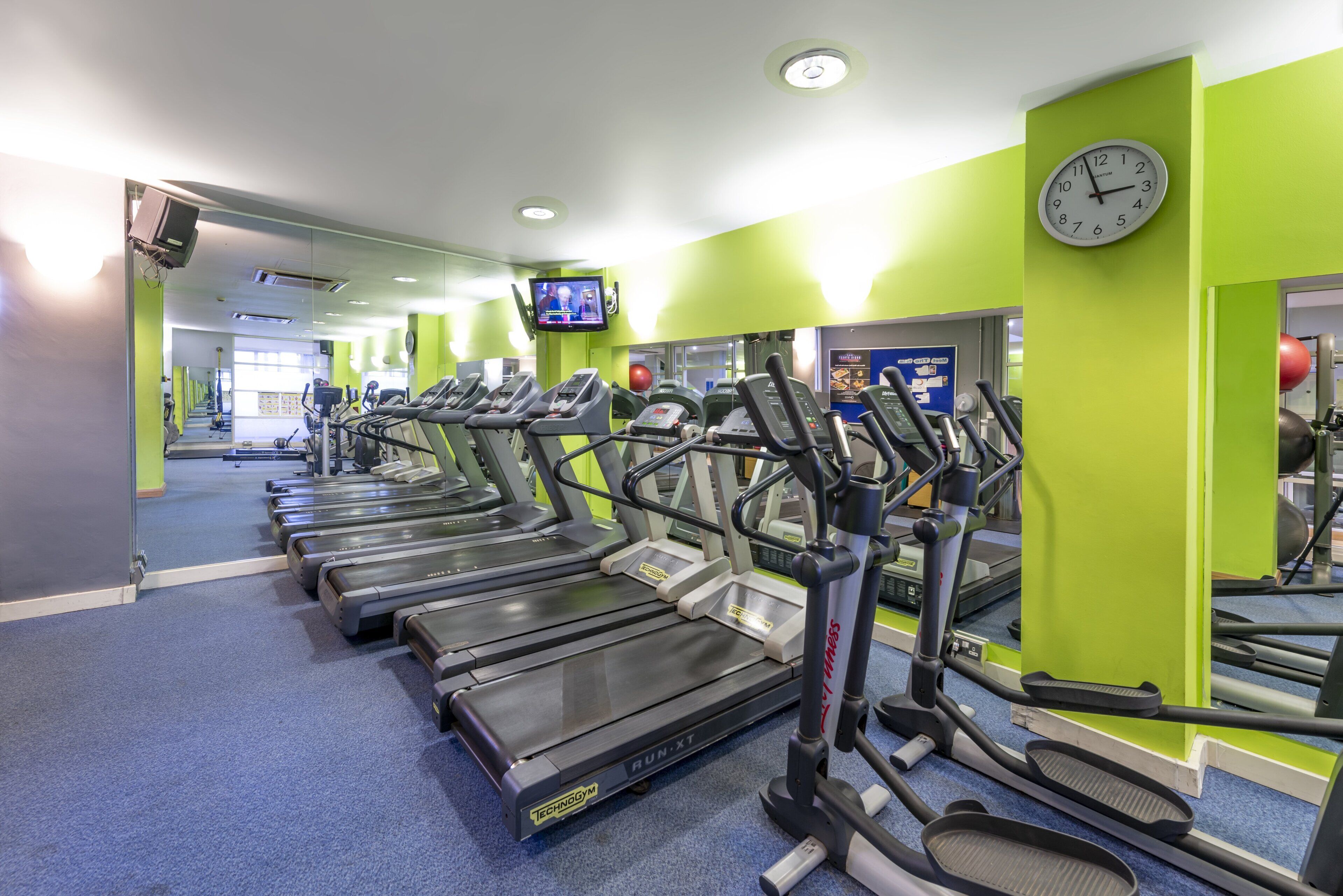 A gym with treadmills lined up against green walls, a clock, and a TV screen.