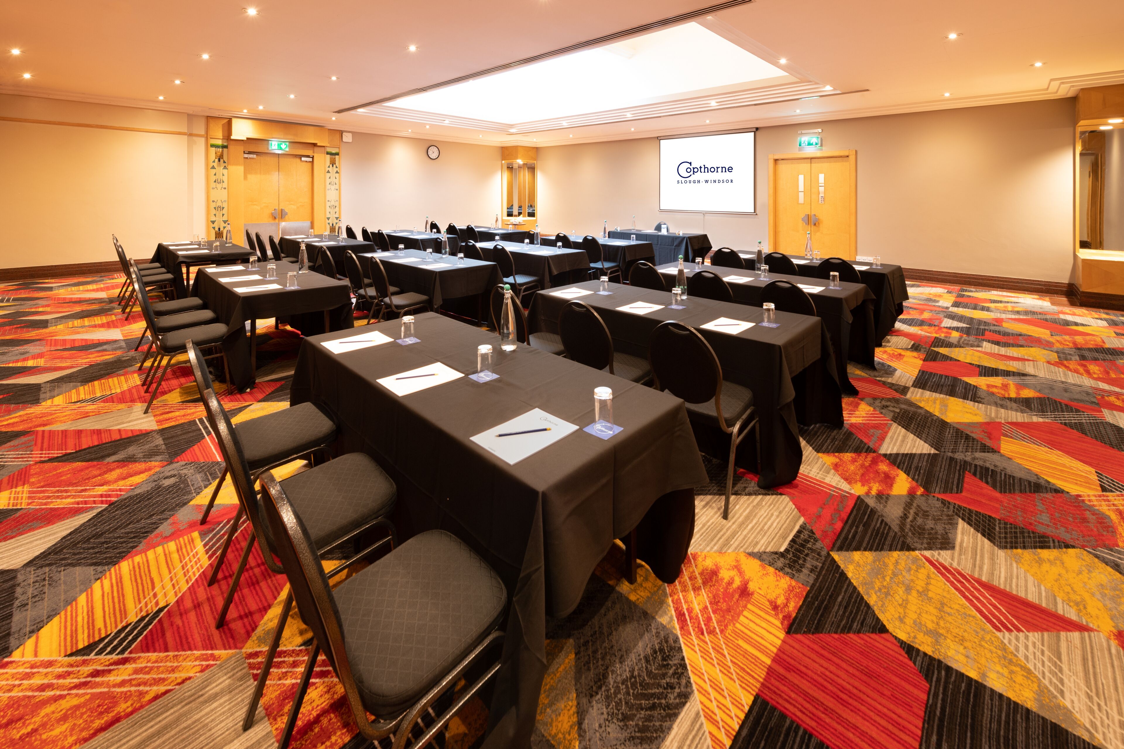 A conference room with multiple tables and chairs, equipped with water bottles and notepads, carpeted floor, and a projection screen displaying the hotel name.