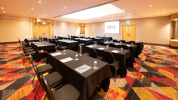 A conference room with multiple tables and chairs, equipped with water bottles and notepads, carpeted floor, and a projection screen displaying the hotel name.