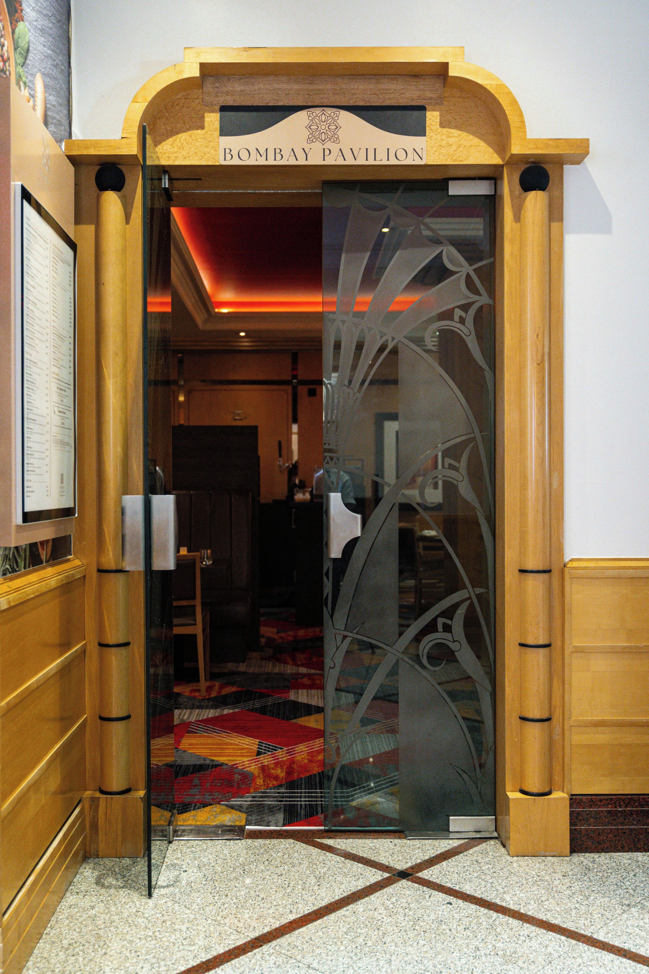Una entrada a un edificio con la leyenda 'Bombay Pavilion', con una puerta de cristal que presenta ornamentación metálica intrincada.