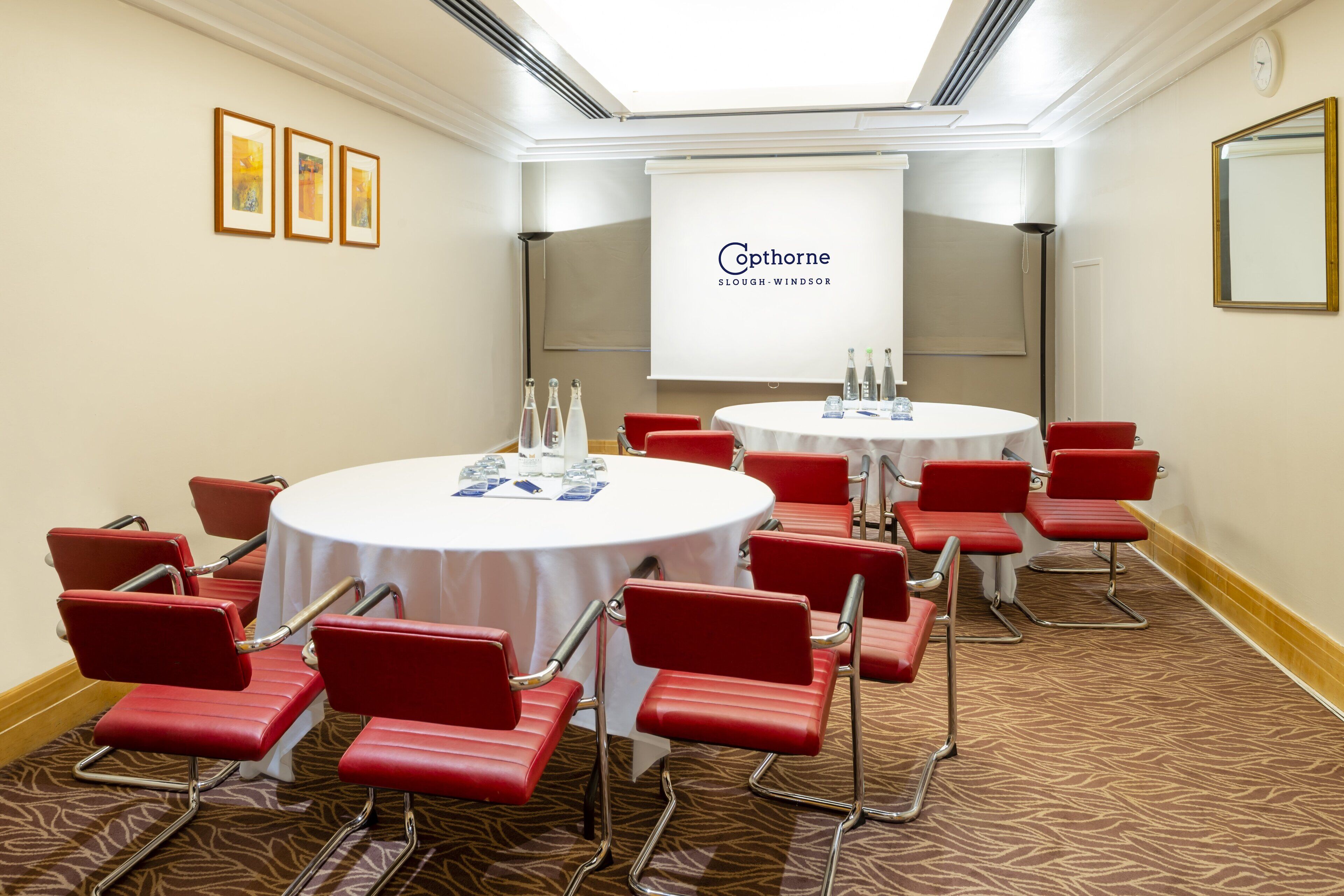 A conference room with two round tables, red chairs, a projector screen, and framed artwork on the wall.