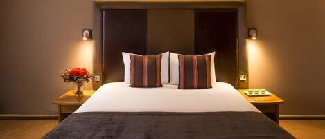 A hotel room with a large bed, two bedside tables, a nightstand, a vase with flowers, and a lamp.