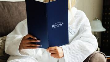 Eine Person hält ein blaues Buch mit dem Schriftzug 'Retrouvé', sitzt auf einem braunen Sofa mit einer weißen Decke. Im Vordergrund liegen ein weißes Tuch und ein Stielglas.