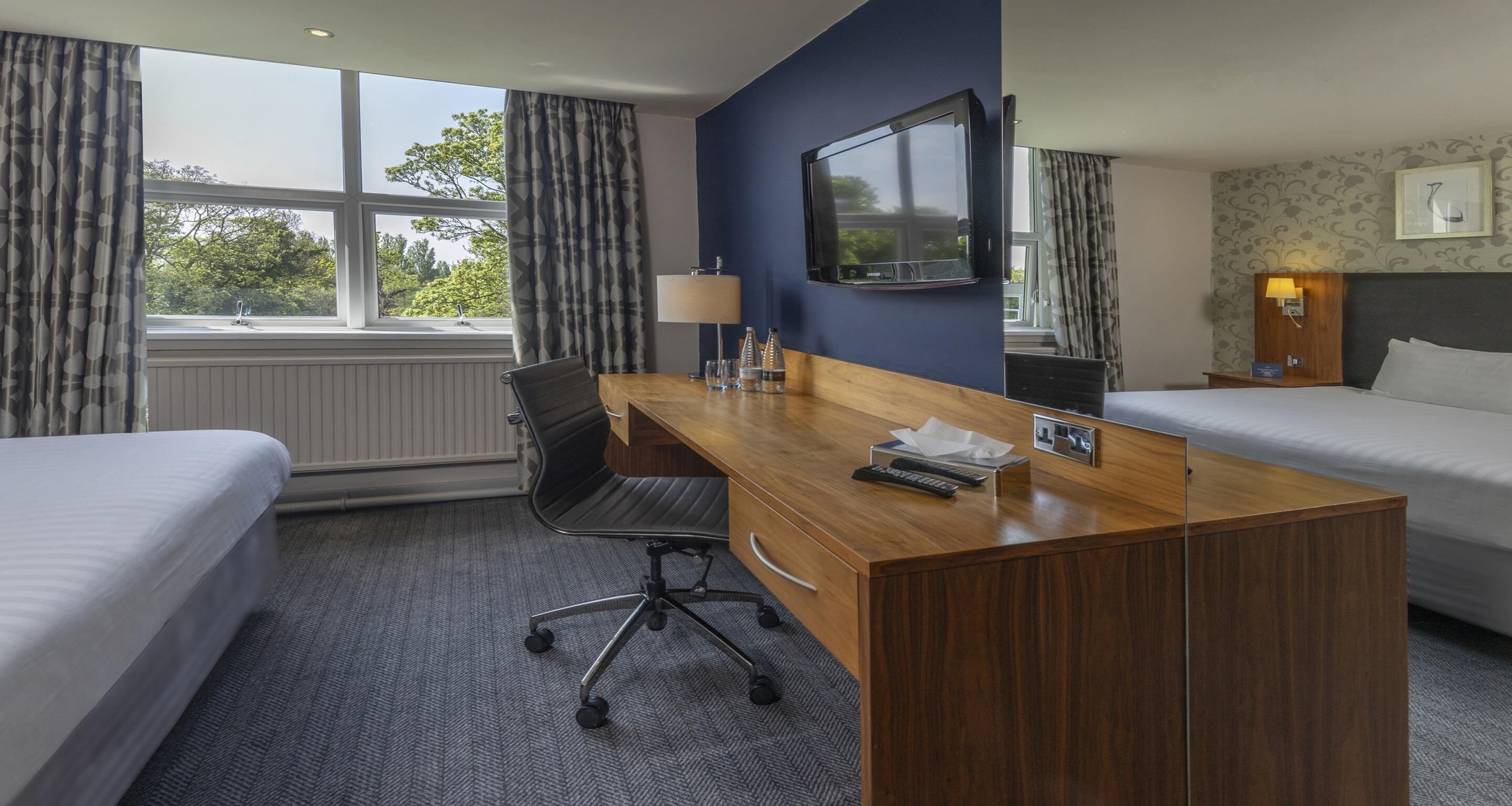 A hotel room with two beds, a wooden desk, a chair, a TV, a lamp, and a nightstand with a lamp.