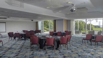 A conference room with round tables and chairs, large windows with a view of greenery and a bridge, and a projector screen on the wall.