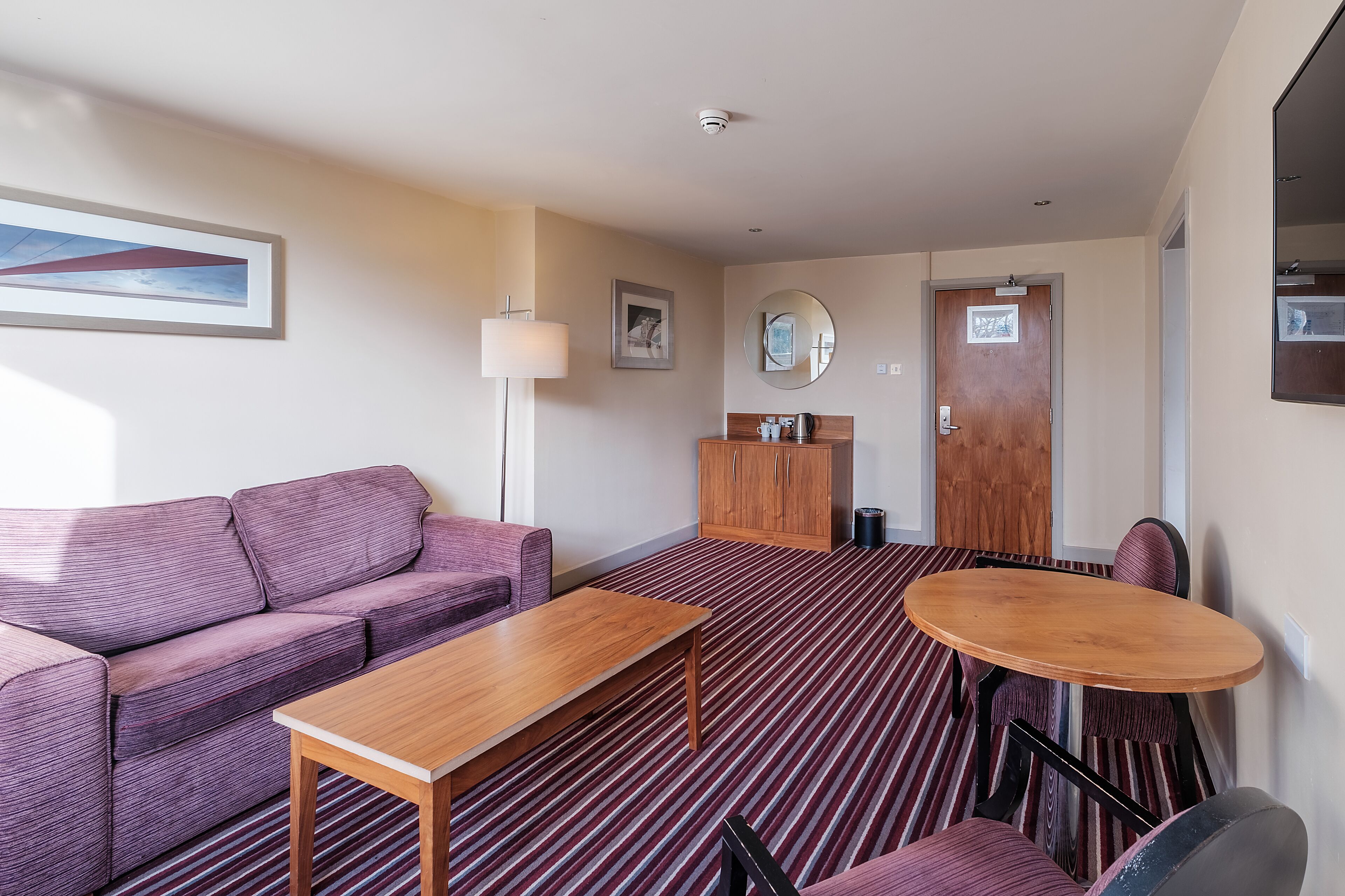A hotel room with a purple sofa, a wooden table, a round table with chairs, a mirror, and a door with a sign.