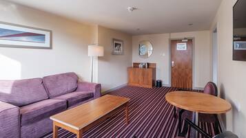 A hotel room with a purple sofa, a wooden table, a round table with chairs, a mirror, and a door with a sign.