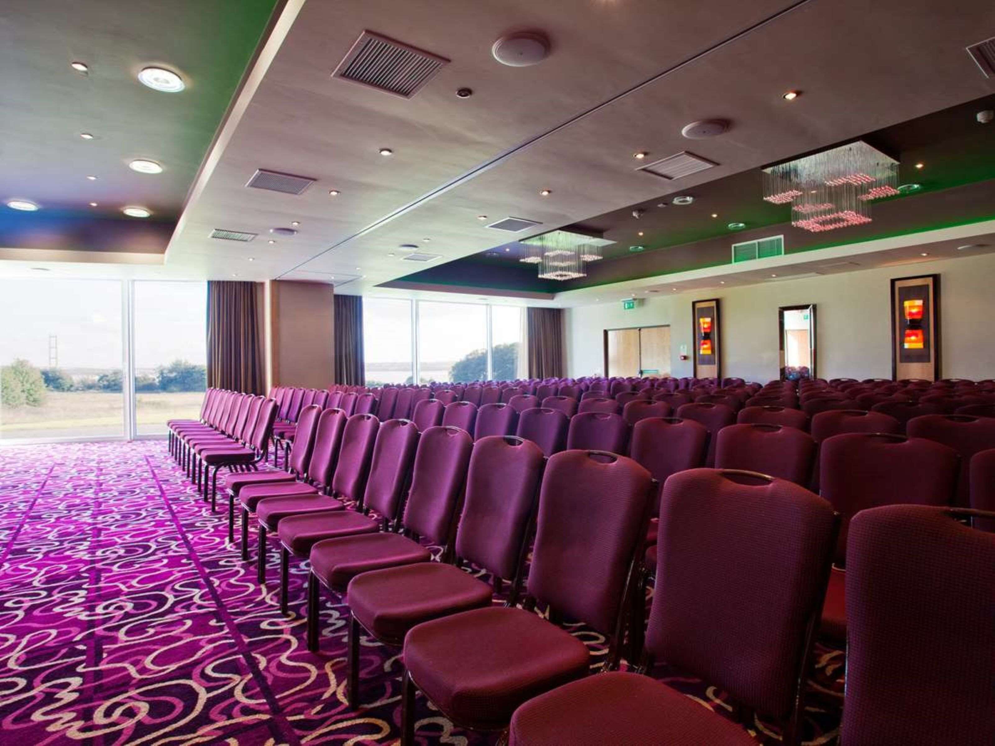 A conference room with rows of purple chairs, a patterned carpet, and large windows offering a view of the outdoors.