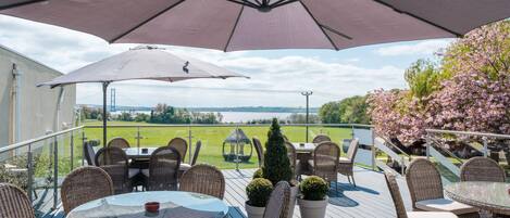 Una terraza con mobiliario de mimbre, un gran paraguas y vistas a un lago y flores de cerezo.