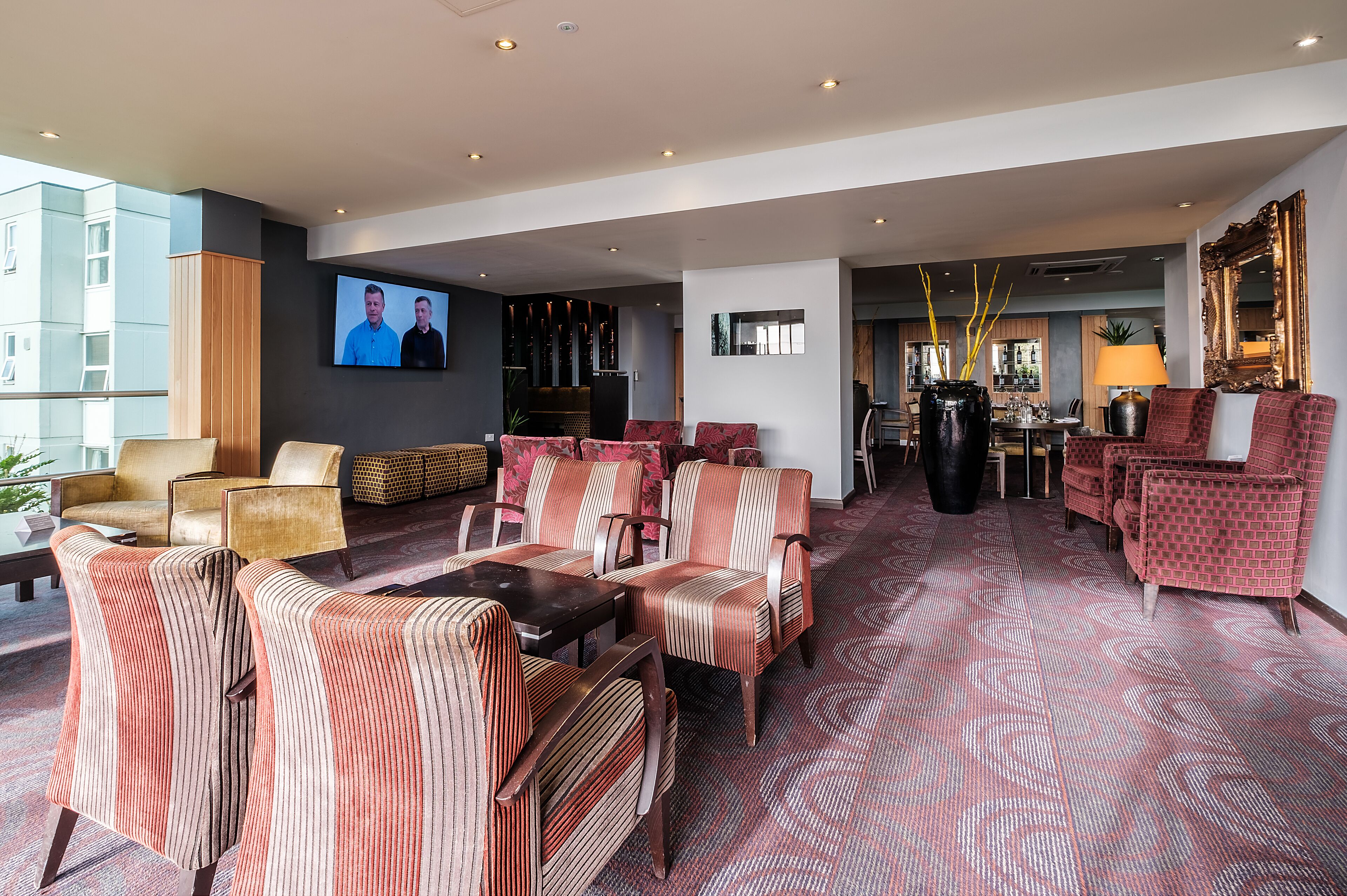 A modern hotel lobby with a seating area, a television screen, and a dining area.