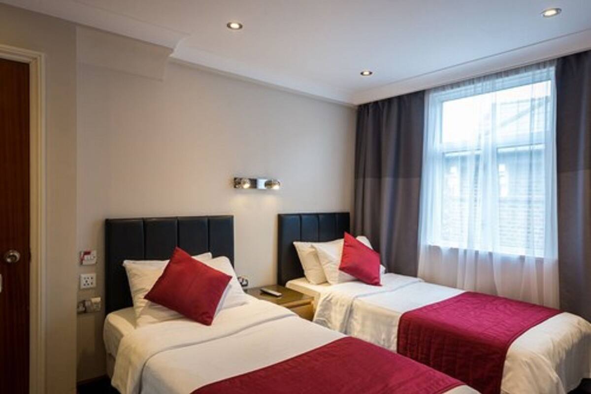 Una camera d'albergo con due letti, ognuno dotato di un cuscino rosso decorativo, un comodino e un punto luce fissato al muro.