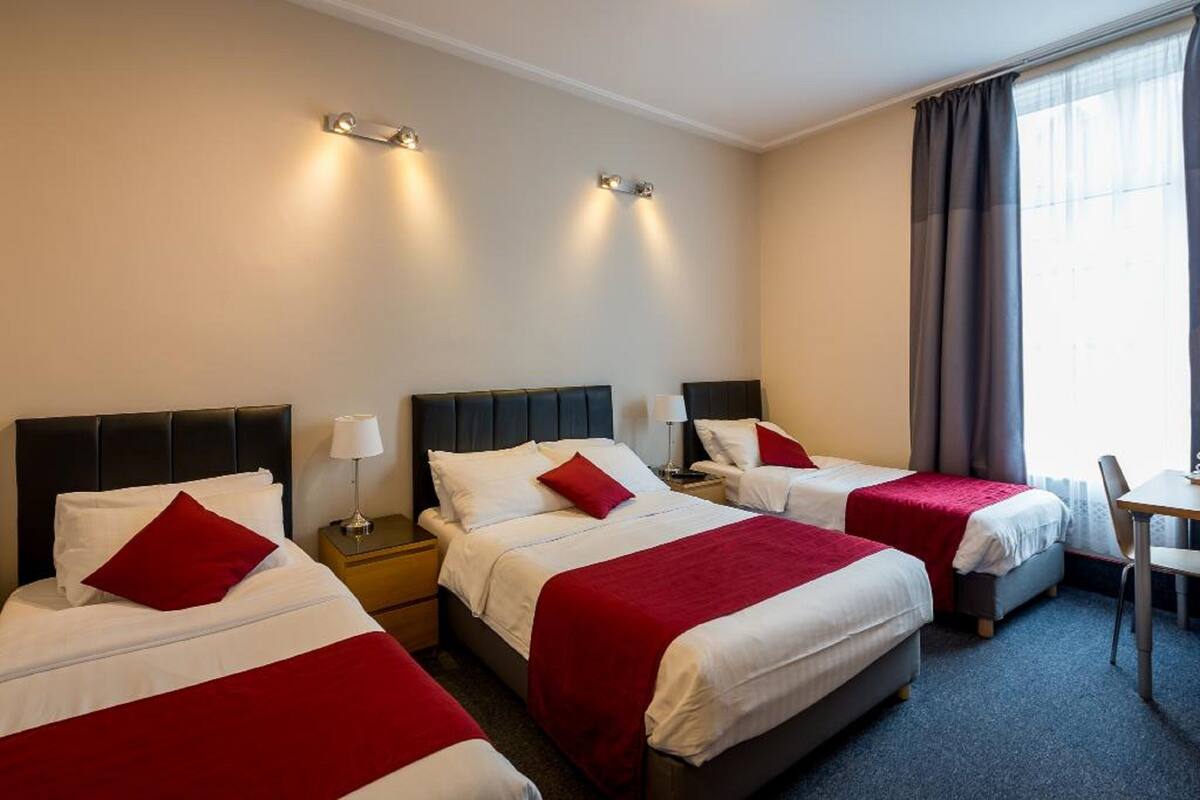 Una camera d'albergo con tre letti, ognuno con copripiumino rosso e bianco, e un tavolino con una sedia.