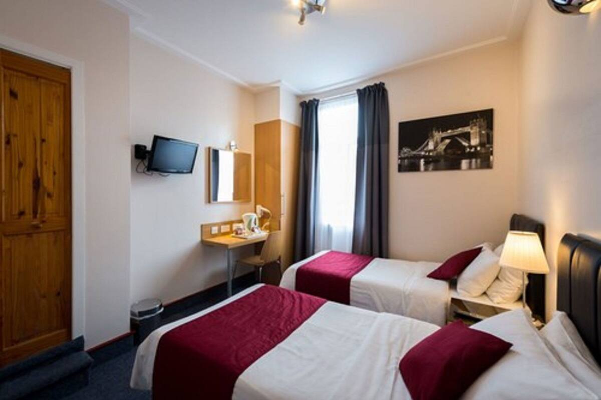 Una camera d'albergo con due letti, una scrivania, un televisore, una finestra con tende e un quadro appeso al muro.