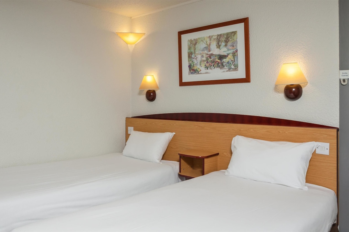 A hotel room with two single beds, a wooden headboard, wall-mounted lamps, and a framed picture on the wall.