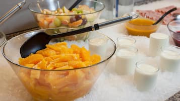 A bowl of orange slices on ice with a spoon, surrounded by small glasses of milk and a bowl of mixed fruit salad.