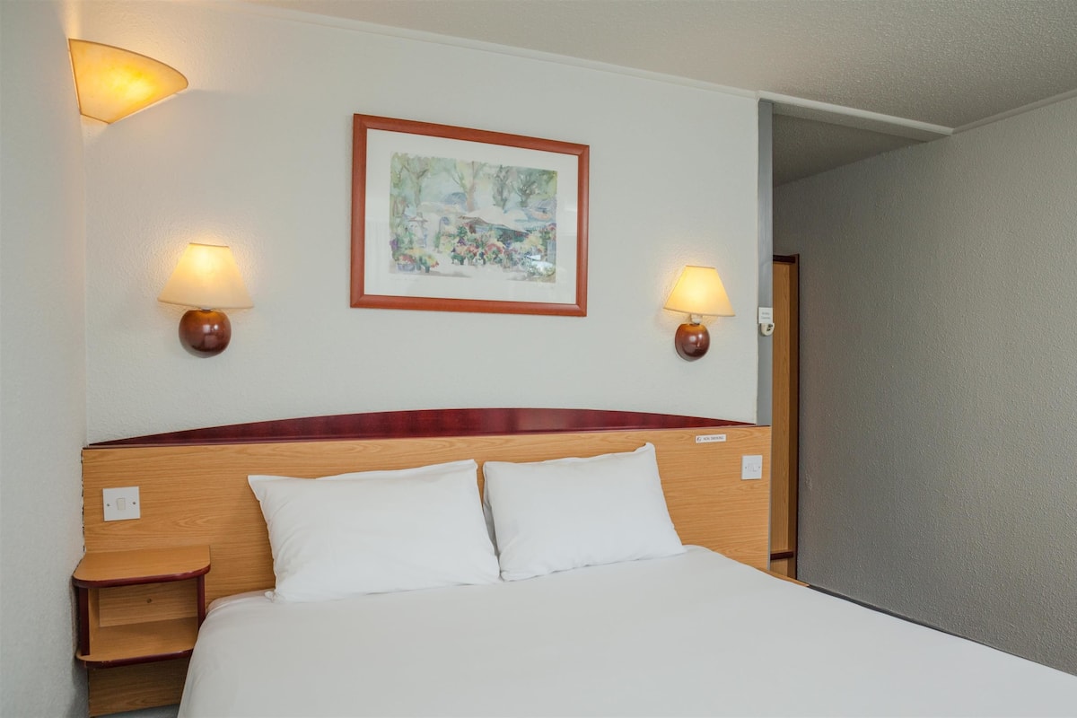 A hotel room with a wooden headboard, two bedside lamps, and a framed painting on the wall.