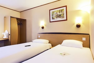 A hotel room with two beds, a wooden headboard, a wall-mounted lamp, a framed picture, and a small table with a remote control.