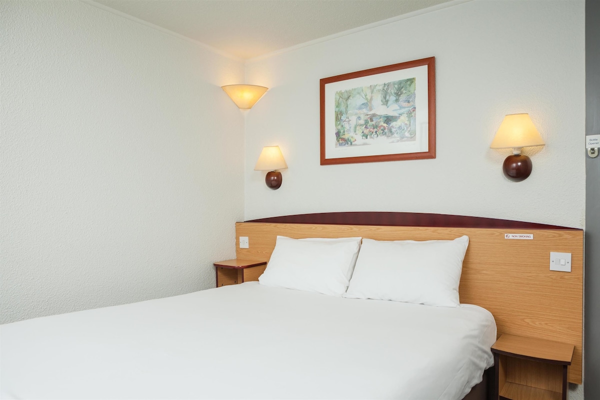 A hotel room with a wooden headboard, two bedside lamps, a framed picture on the wall, and a sign indicating a non-smoking area.