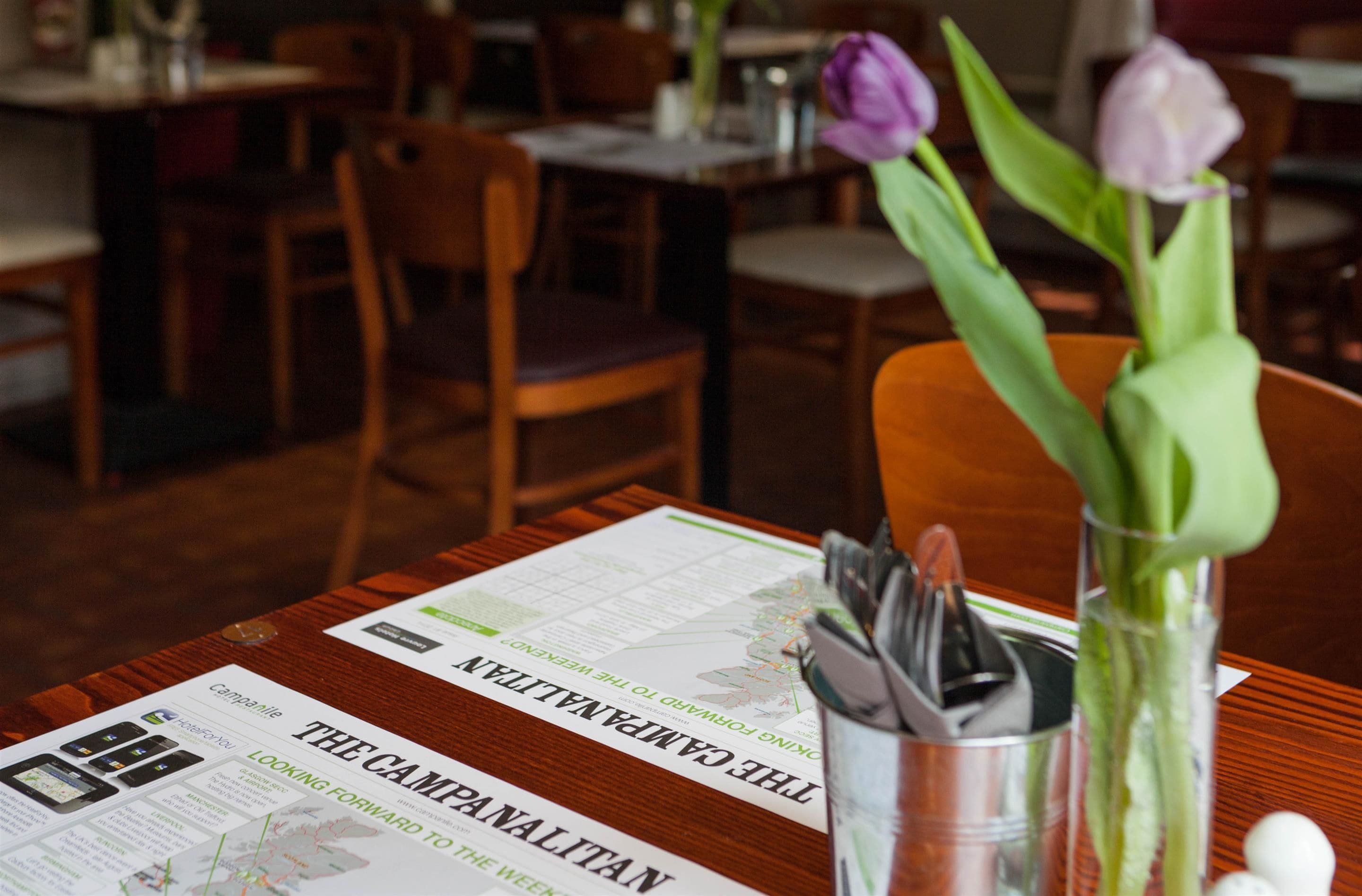A restaurant table with a menu, a metal utensil holder, and two tulips in a vase.