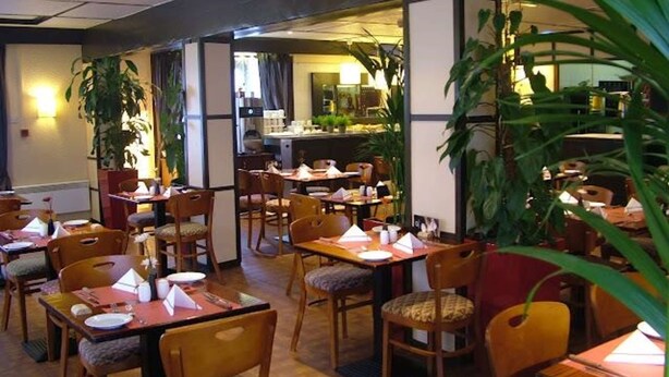 Un restaurant avec des tables et des chaises en bois, dont certaines sont dotées de coussins. Des plantes décorent la pièce, et on aperçoit un espace cuisine au fond.