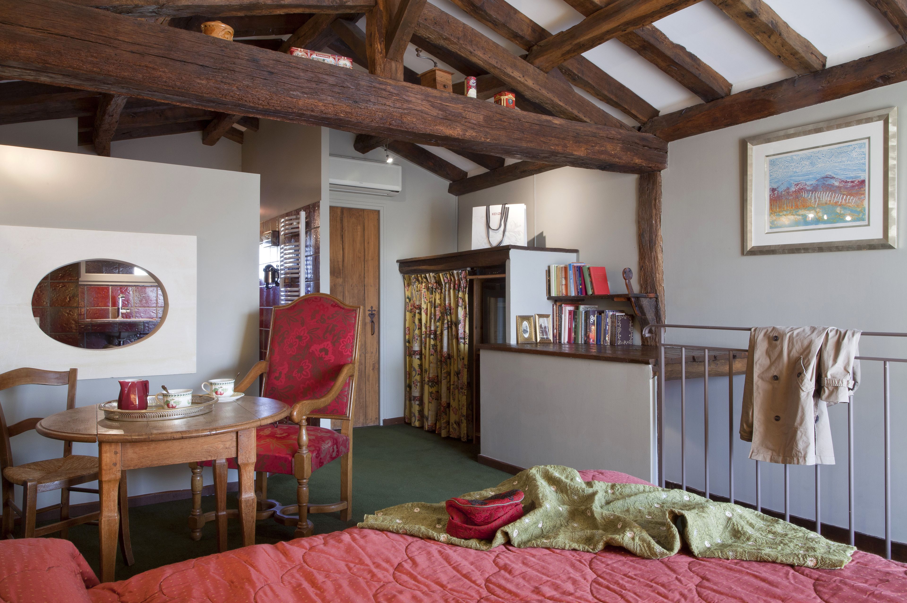 A cozy room with a wooden ceiling, a bed with a red quilt, a wooden table with a pitcher and cups, a red chair, and a bookshelf with books and a framed picture.