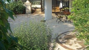 A traditional villa with a tiled roof, a covered patio with a table and chairs, and lush greenery surrounding the property.