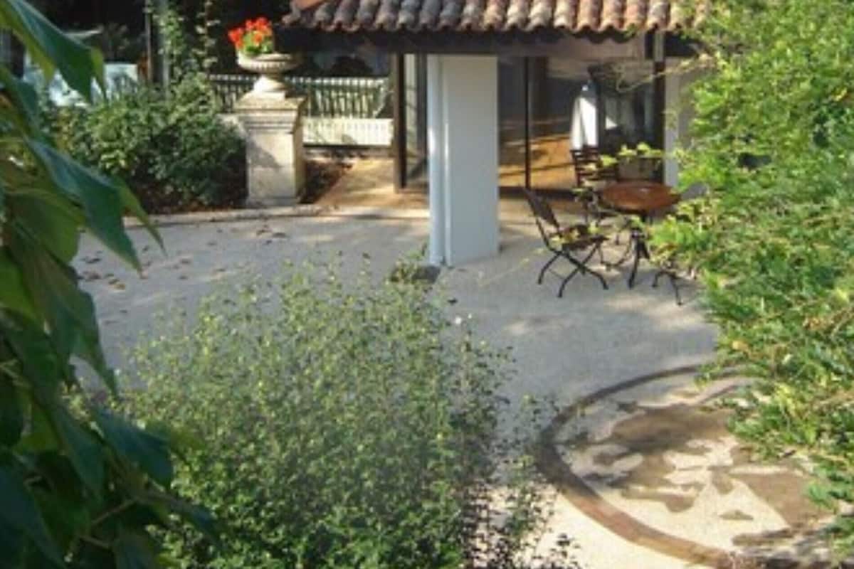 Eine traditionelle Villa mit Ziegeldach, einer überdachten Terrasse mit Tisch und Stühlen und üppiger Vegetation rund um das Anwesen.
