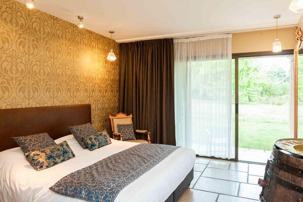 A hotel room with a large bed, a wooden headboard, patterned curtains, and a view of a garden through a sliding door.