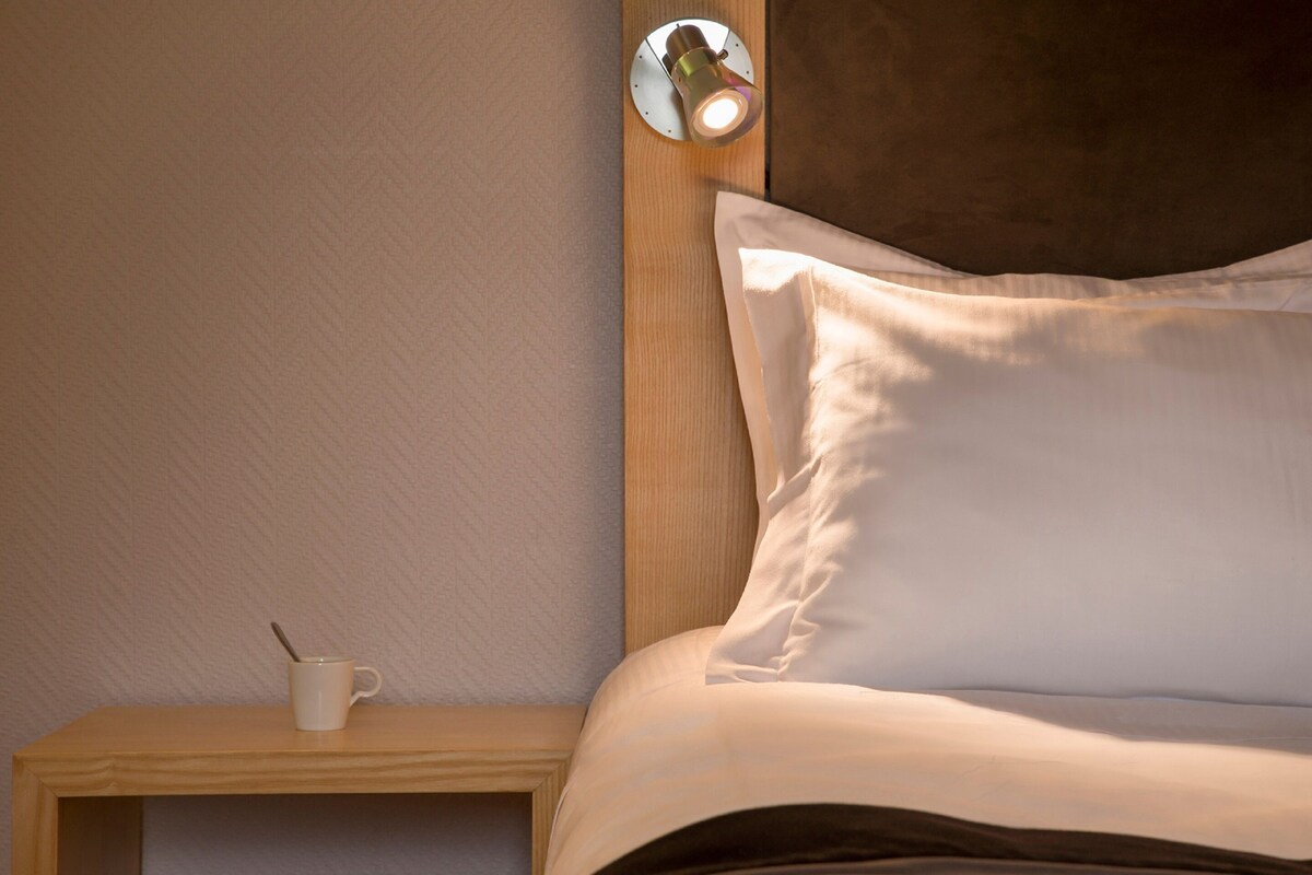 Una cama bien hecha con un almohadón, una mesita de noche con una taza y una cuchara, y una lámpara fijada a la pared.