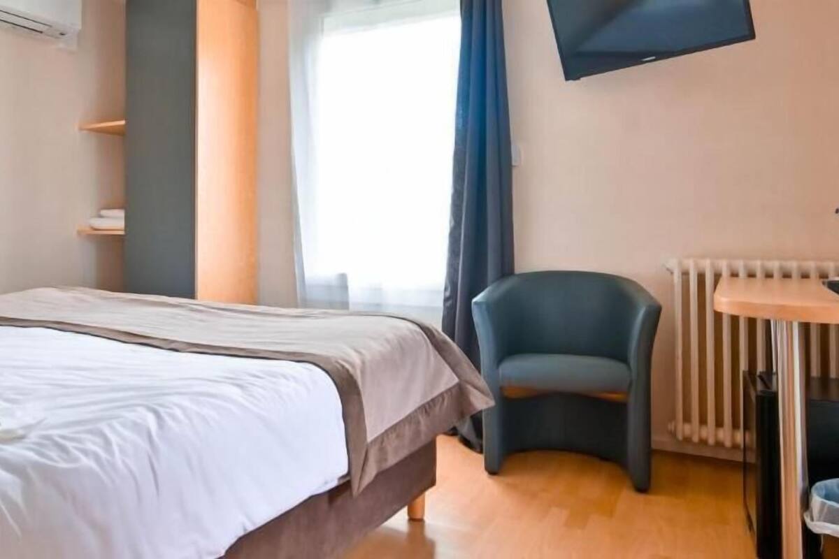 A hotel room with a bed, a chair, a small table, a TV, and a window with curtains.