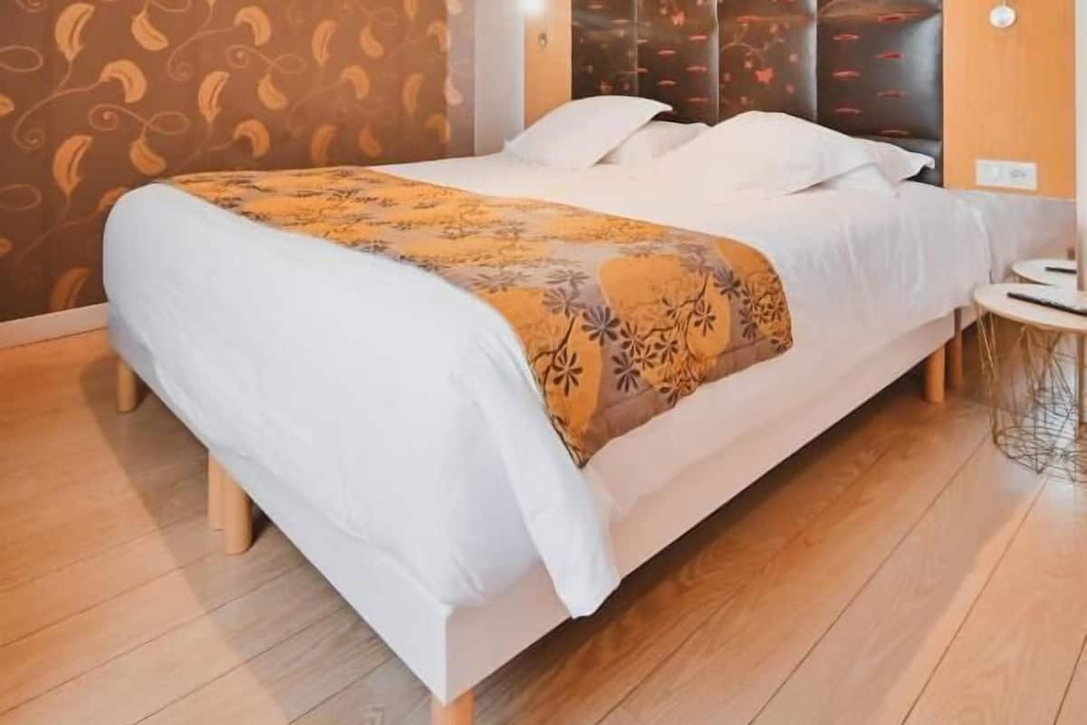 A modern hotel room with a large bed, wooden flooring, a patterned wall, and a decorative headboard.