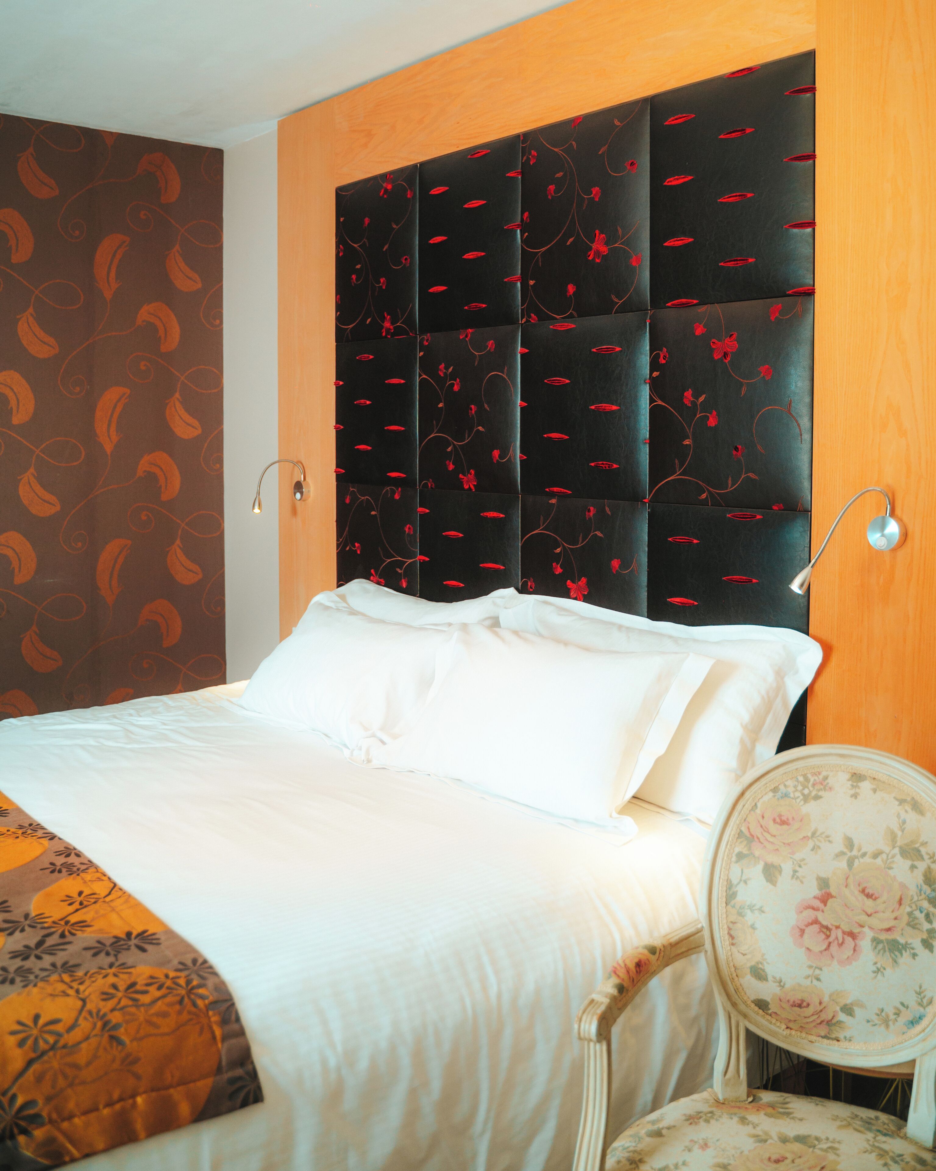 Una cama bien hecha con sábanas blancas, cabecera oscura con motivos florales rojos y una silla estampada con flores.