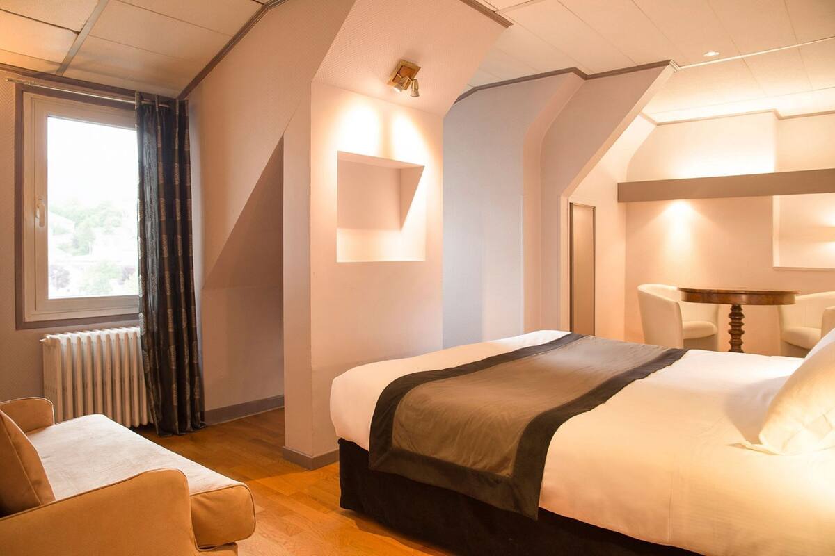 A hotel room with a bed, a chair, a table, a radiator, and a window with curtains.