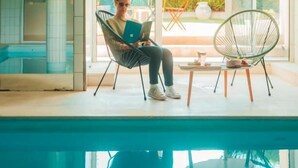 Eine Person sitzt an einem Pool auf einem Stuhl mit einem Laptop, ein Stuhl mit Sitzfläche aus Korbgeflecht, und Blick ins Grüne.