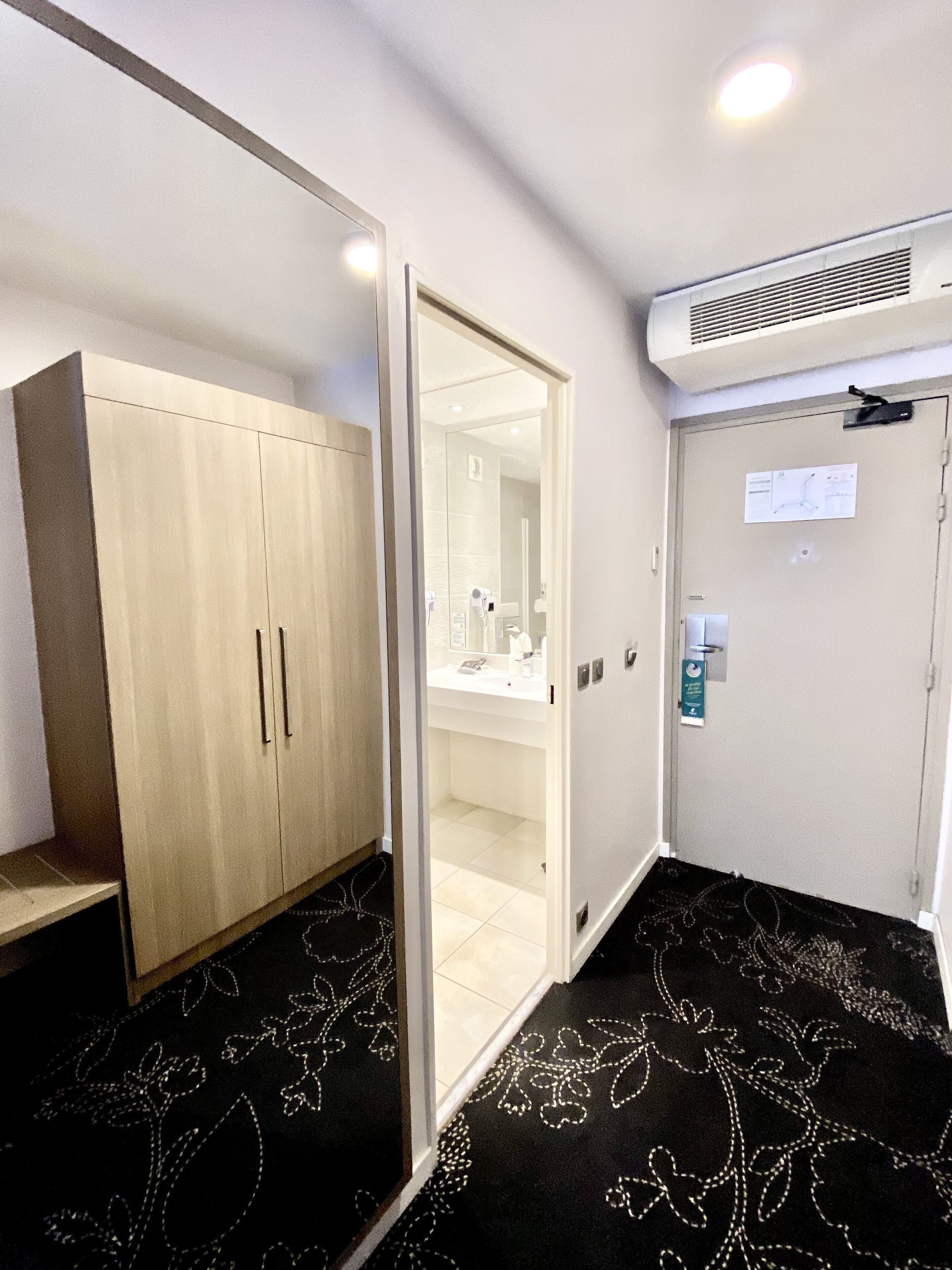 Una habitación de hotel con un armario, un baño con espejo y una alfombra estampada con motivos florales.