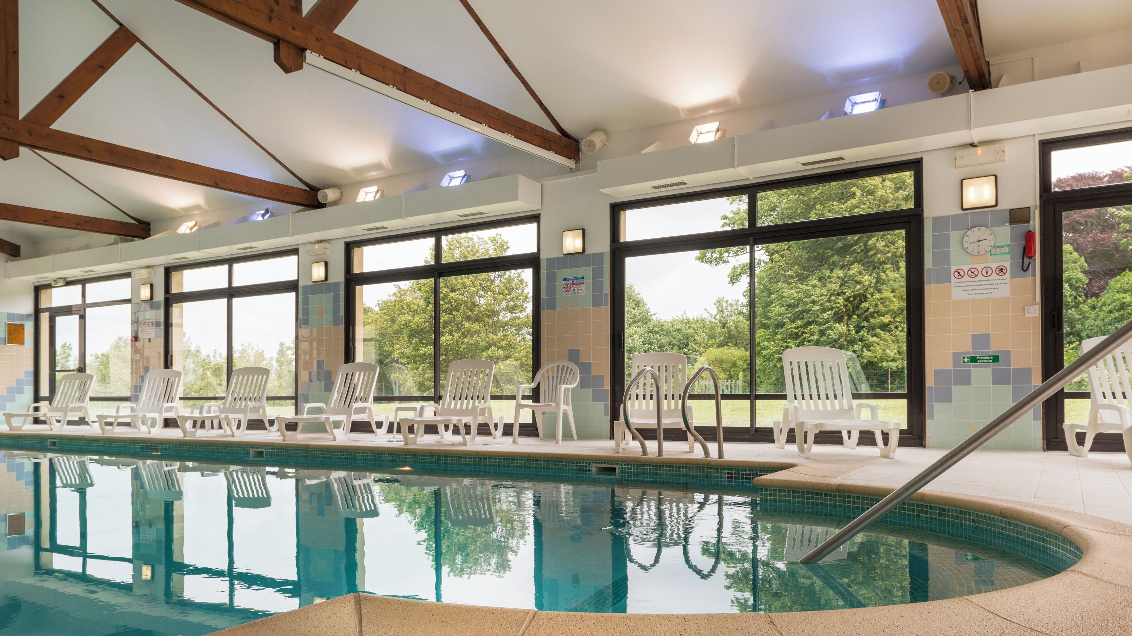 An indoor swimming pool with clear glass walls, wooden beams, and white lounge chairs.