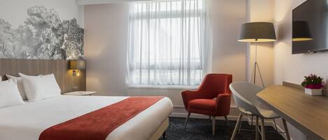 Hotellihuone, jossa on suuri sänky, työpöytä, tuoli, lamppu, televisio ja ikkuna, jossa on ohuet verhot.