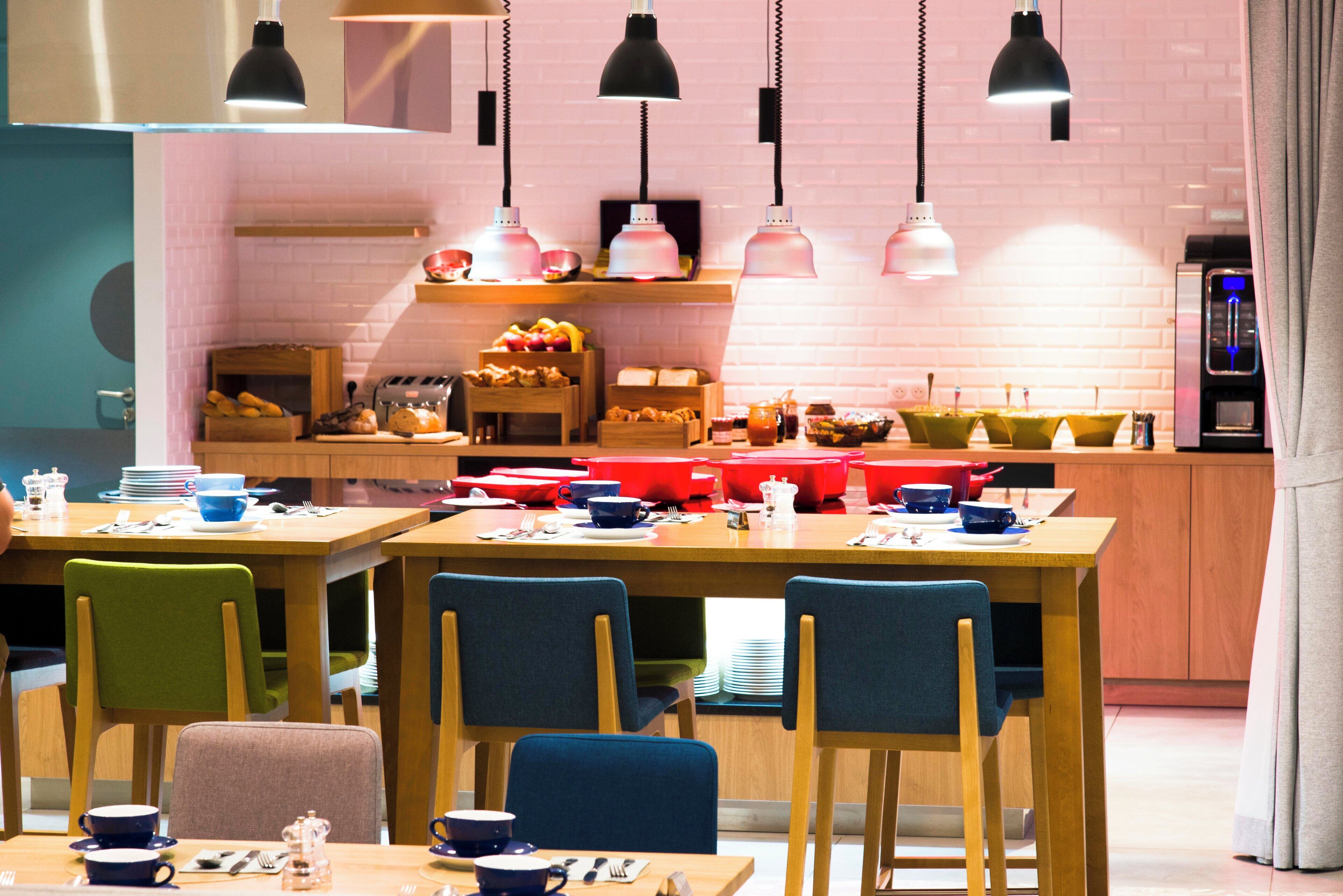 A modern dining area with wooden tables and chairs, a kitchen in the background, and pendant lighting.