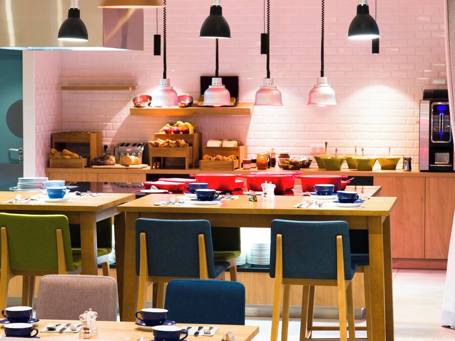Uma área de refeições moderna com mesas e cadeiras de madeira, uma cozinha ao fundo e iluminação pendente.