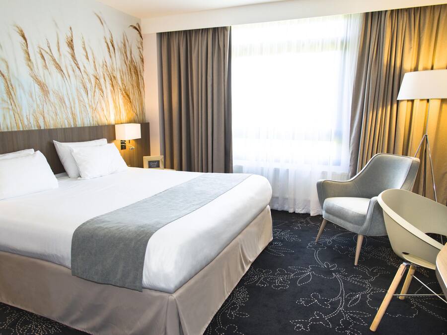Una habitación de hotel con una cama grande, una silla, una lámpara y un ventanal con cortinas.