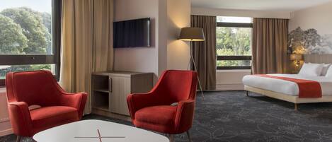 A hotel room with a bed, two red chairs, a desk, a TV, and large windows with curtains.