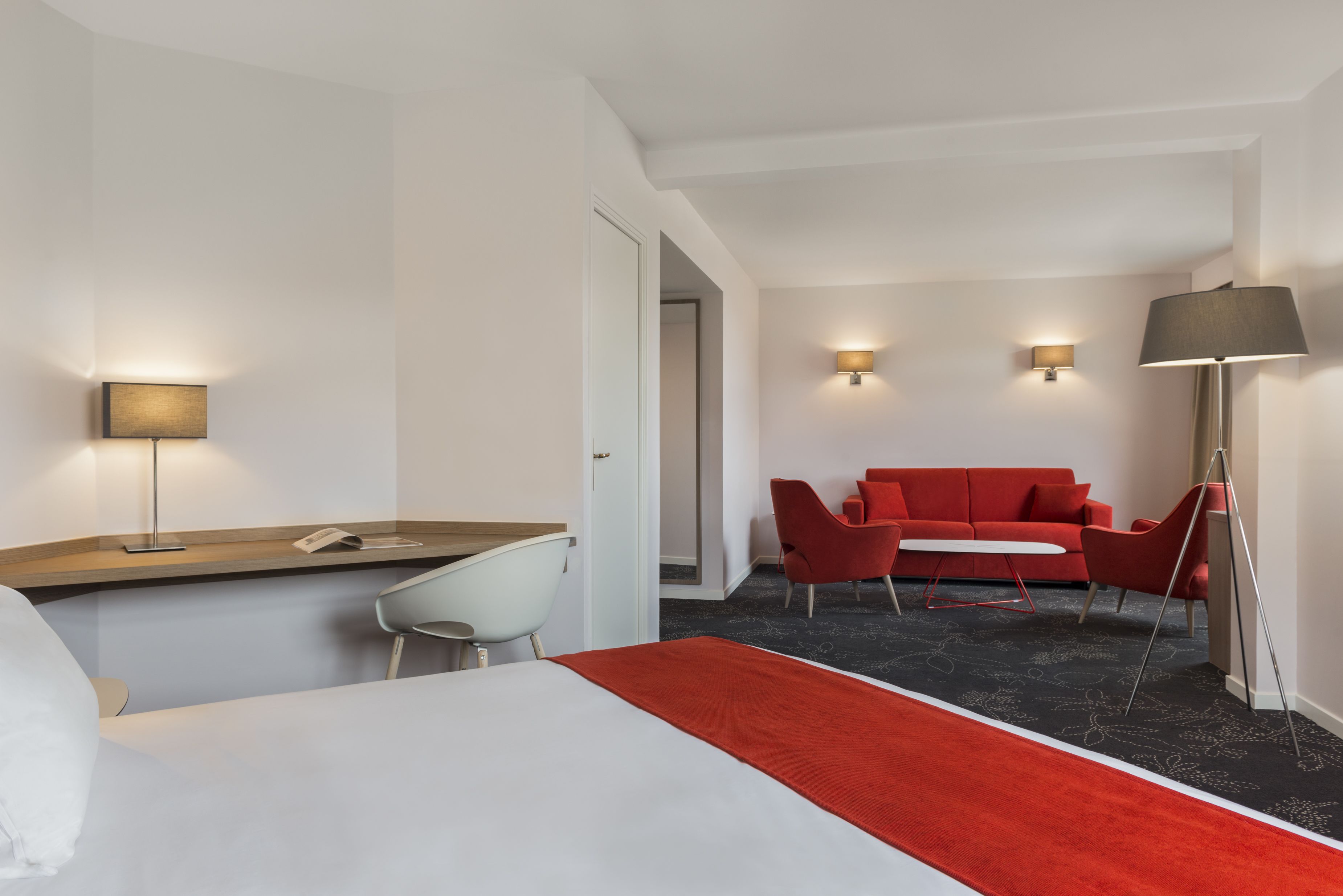 Ett hotellrum med en säng, ett skrivbord, en stol, en röd soffa och två vägglampor.