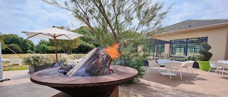 Una estructura exterior para fuego con leña, rodeada de muebles de jardín y un edificio con ventanas amplias.