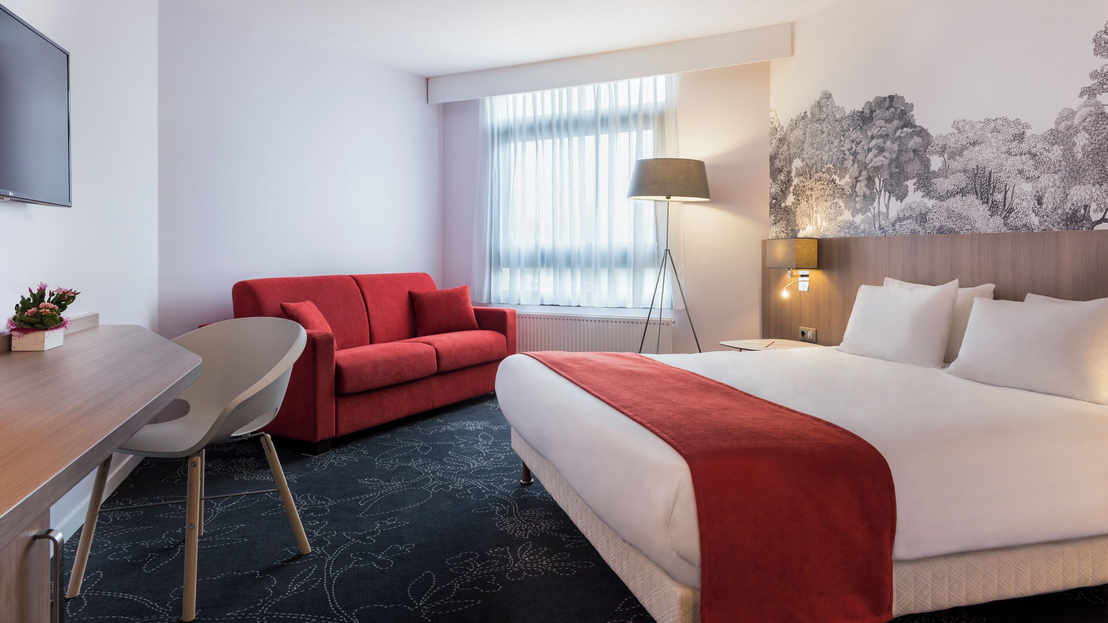 A hotel room with a bed, a red sofa, a desk with a chair, a lamp, and a large window with sheer curtains.