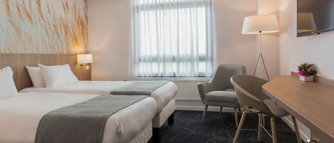 Habitación de hotel con dos camas, un escritorio, una silla, una lámpara y un ventanal con cortinas translúcidas.