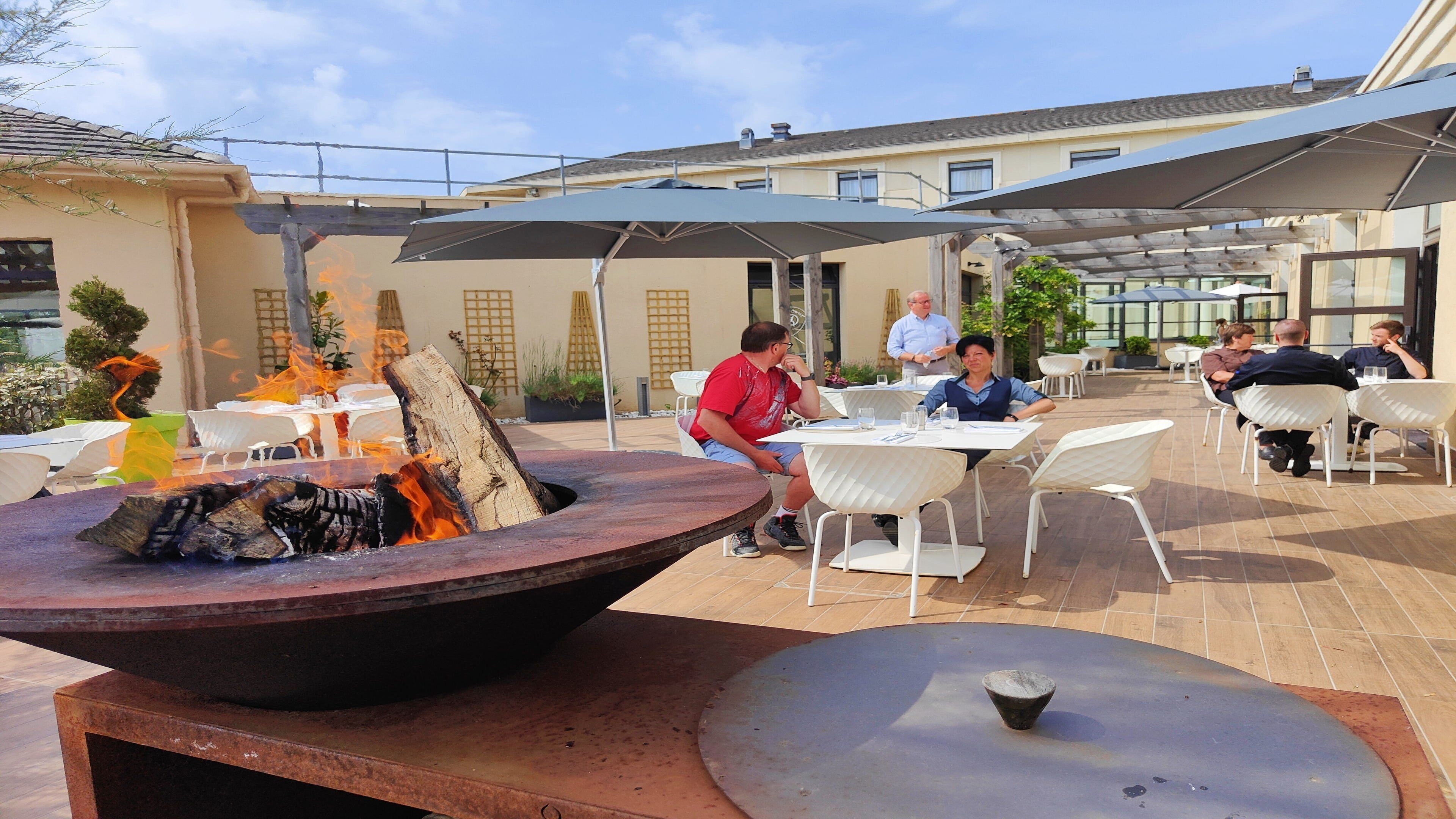 Un espace extérieur aménagé pour les repas, avec des tables et des chaises, un foyer et des parasols pour créer de l’ombre.