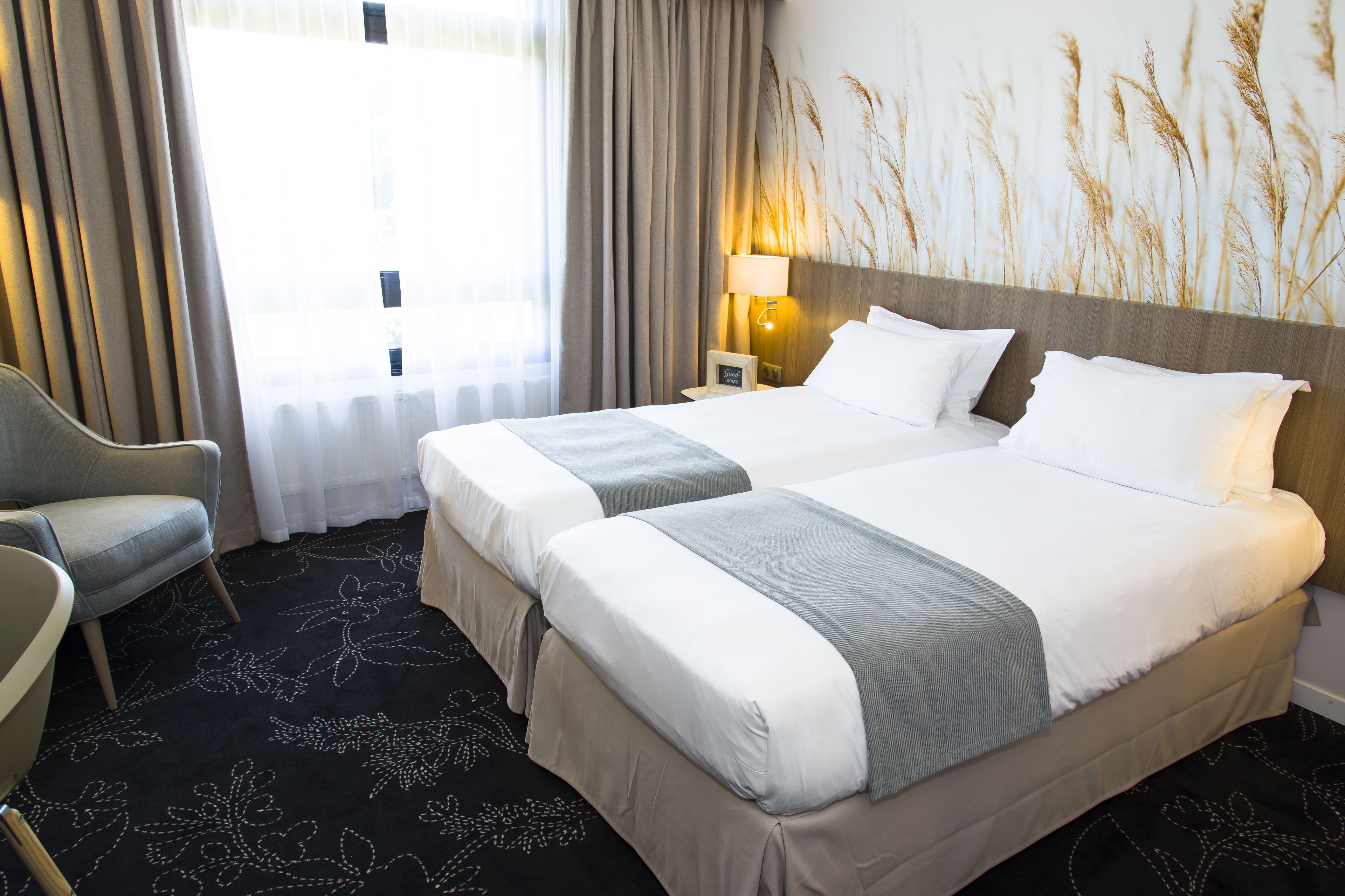 Habitación de hotel con dos camas, una silla, una lámpara y un ventanal grande con cortinas.