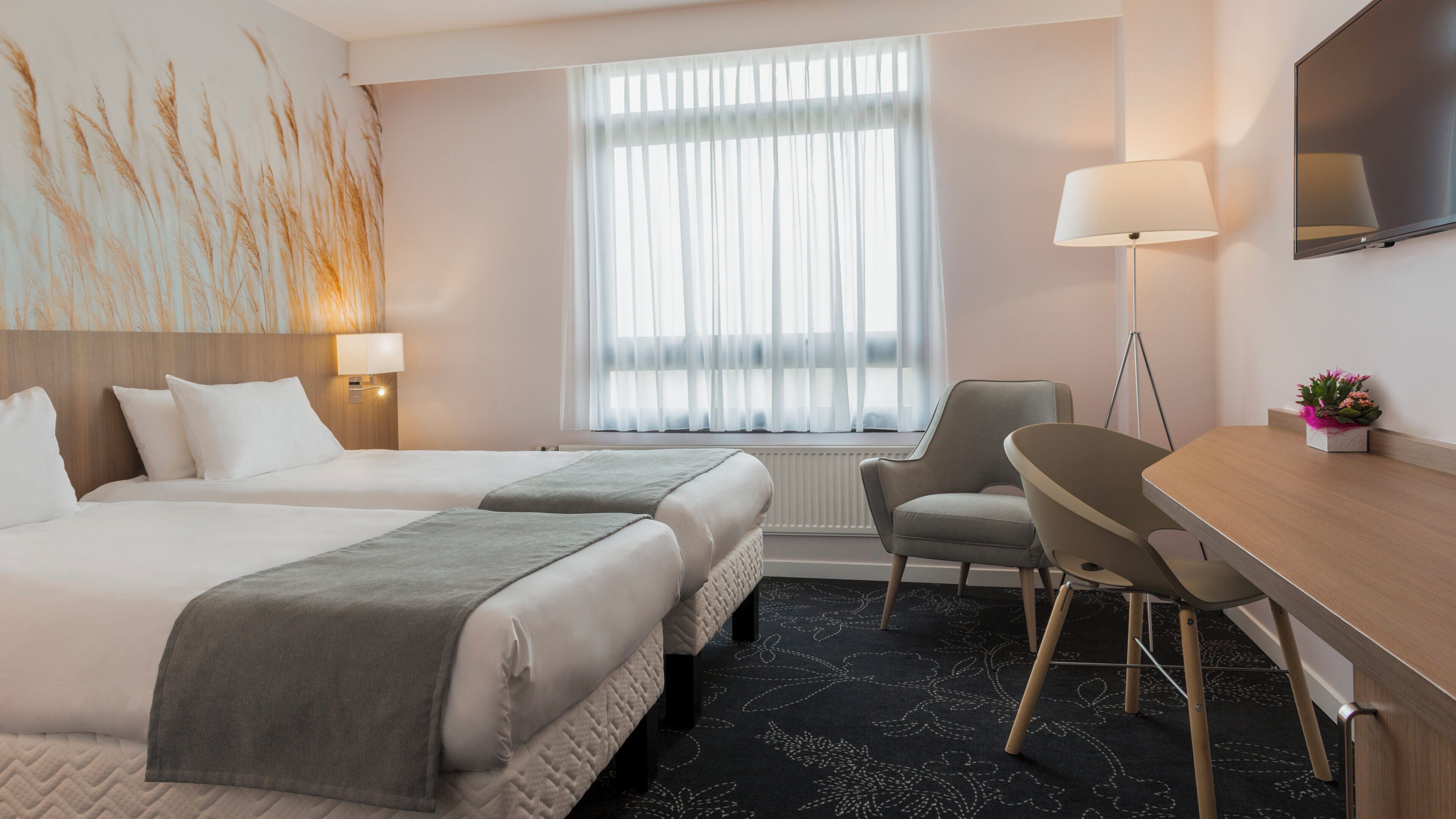 A hotel room with two beds, a desk, a chair, a lamp, and a large window with sheer curtains.